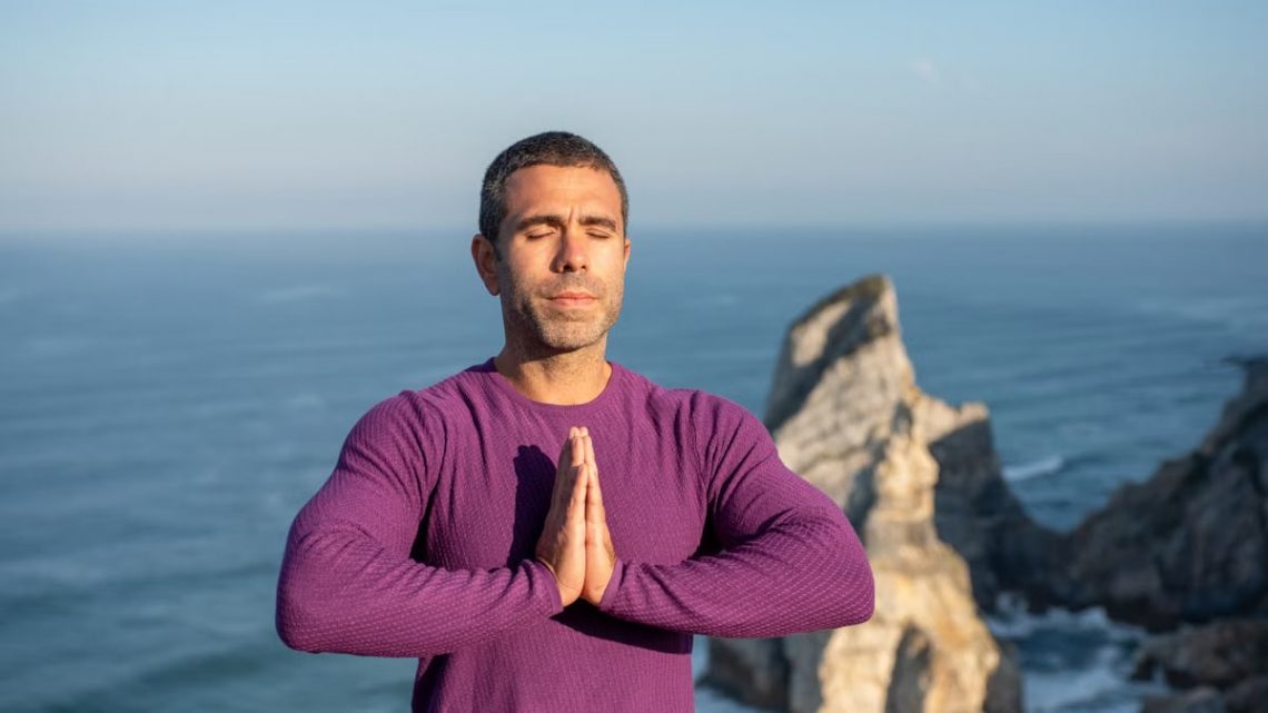Man meditating with hands together by a rocky coastline in Portugal, ocean in background.