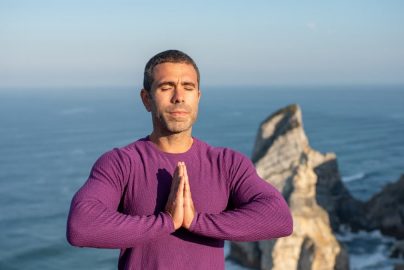 Man meditating with hands together by a rocky coastline in Portugal, ocean in background.