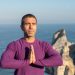 Man meditating with hands together by a rocky coastline in Portugal, ocean in background.