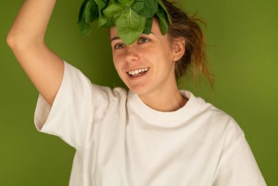 Cheerful adult female in white t shirt with fresh spinach foliage against forehead looking up