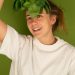Cheerful adult female in white t shirt with fresh spinach foliage against forehead looking up